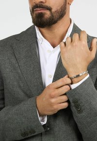 Bracciale d'argento con una finitura liscia e lucida. Indossato su un polso parzialmente visibile, abbinato a un giacca da abito a quadri grigi e una camicia bianca.