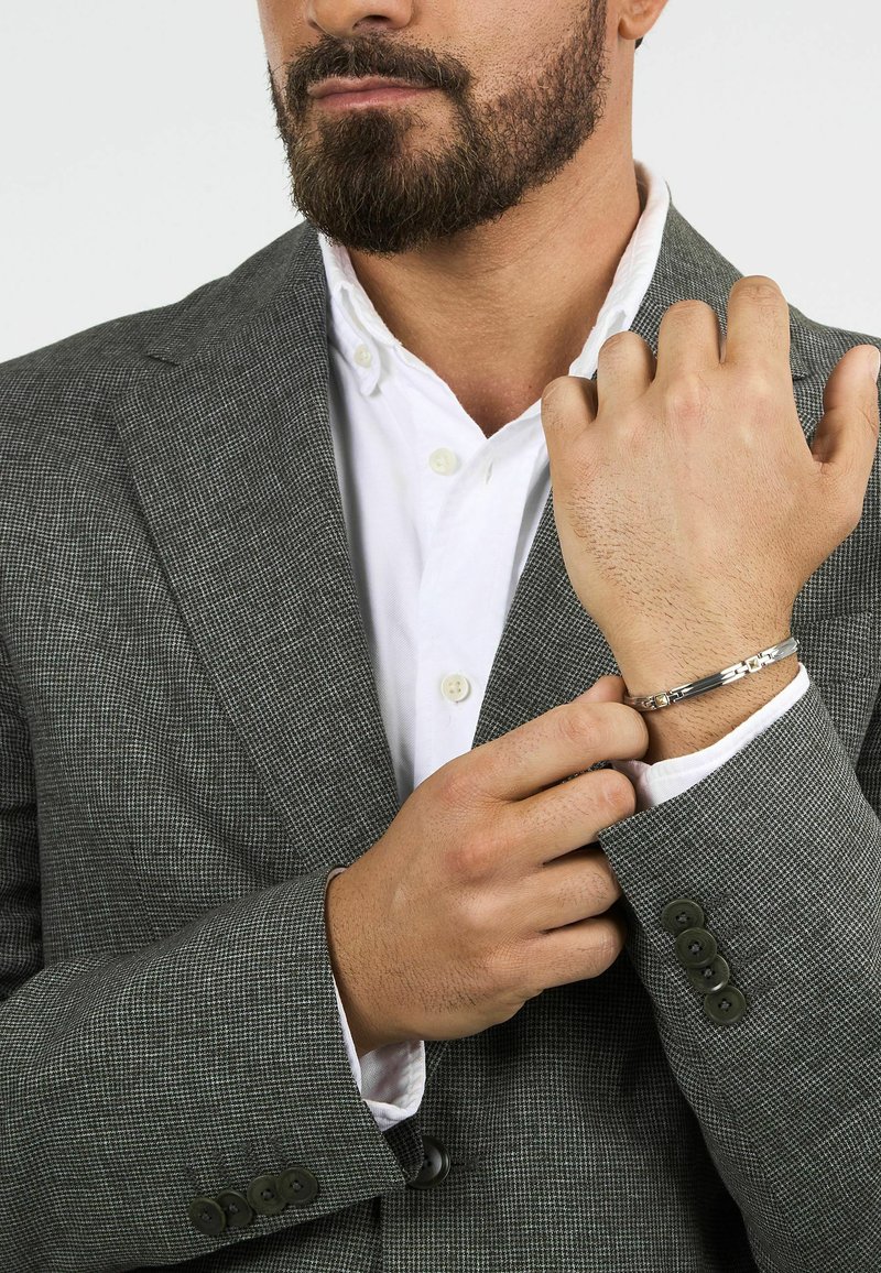 Bracciale d'argento con una finitura liscia e lucida. Indossato su un polso parzialmente visibile, abbinato a un giacca da abito a quadri grigi e una camicia bianca.