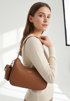 Femme portant un pull crème tient un sac à main en cuir marron Diana&Co Firenze sur l'épaule dans une pièce à l'éclairage doux.