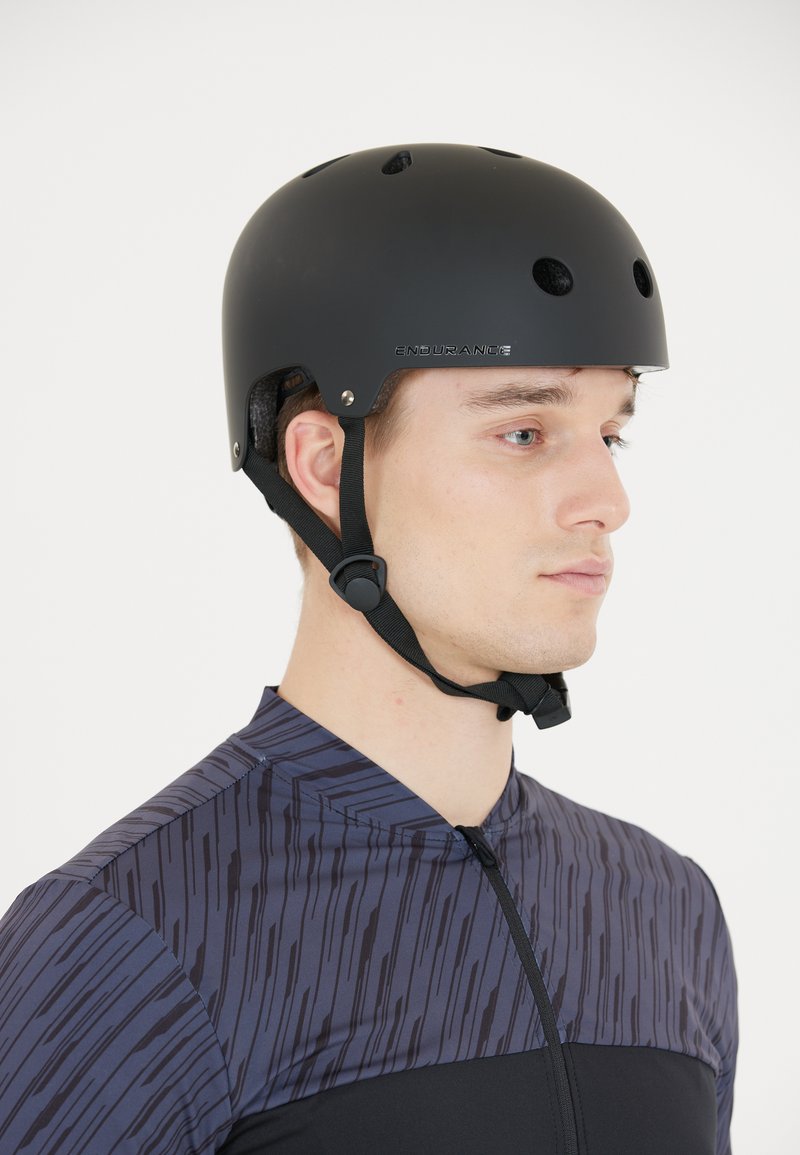 Svart sportshjälm med ventilationshål, justerbara remmar och en matt yta. Bärs av en person i en mönstrad cykeltröja.