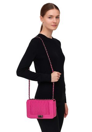Frau mit heller Haut und braunen Haaren trägt ein schwarzes Langarmshirt und hält eine auffällige pink gemusterte Schultertasche mit Kettenriemen.