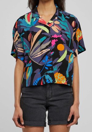 Femme portant une chemise colorée à manches courtes avec des motifs abstraits de feuilles et de fruits, assortie à un short en denim noir retroussé.