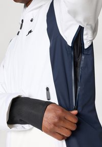 Balta ir tamsiai mėlyna striukė su užtrauktuko kišene ir tinklelio pamušalu, turinti juodą rankovę. Audinys atrodo lygus, su šiek tiek blizgiu paviršiumi.