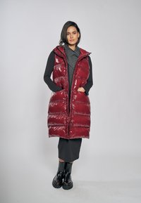 Bordowa kamizelka puchowa o błyszczącym wykończeniu, zapinana na suwak z przodu, z kapturem i bocznymi kieszeniami, noszona na ciemnym swetrze i długiej spódnicy.