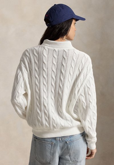 Frau, die einen weißen Strickpullover mit Zopfmuster, hellblaue Jeans und eine marineblaue Baseballkappe trägt, steht mit dem Rücken zur Kamera vor einem neutralen Hintergrund.
