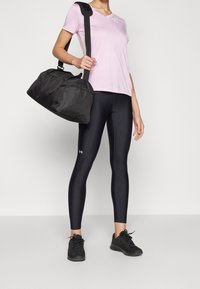 Lichtroze V-hals t-shirt, zwarte leggings met logo, zwarte sporttas en zwarte sportschoenen, allemaal tegen een effen achtergrond.