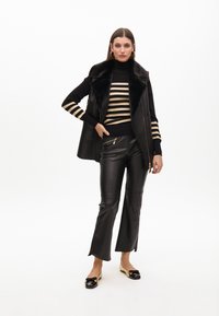 Chaleco de cuero negro con cuello de piel sintética, combinado con un suéter a rayas negras y doradas y pantalones de cuero ajustados negros. Zapatos planos con detalles.