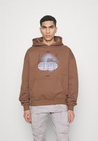 Felpa marrone in tessuto morbido, con un design grafico frontale che include testo e un motivo circolare, e una tasca a marsupio. Abbinata a pantaloni cargo chiari.