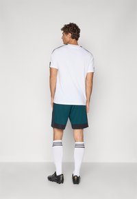 Camiseta deportiva blanca con detalles en negro, combinada con pantalones cortos en color azul verdoso que tienen un dobladillo gris oscuro, calcetas hasta la rodilla en blanco y botas de fútbol negras.