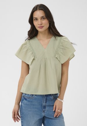 Femme aux longs cheveux foncés portant un chemisier vert clair à col en V avec des manches à volants et un jean bleu en denim, debout devant un fond uni.