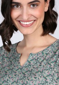 Bluse mit Blumenmuster in Grün und Pink, mit einem Ausschnitt und strukturiertem Stoff. Model mit kurzen, welligen Haaren lächelt in die Kamera.