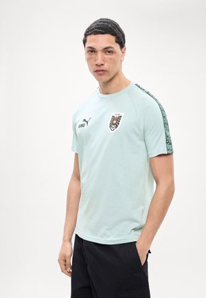 Νέος άνδρας με κοντά σκούρα μαλλιά και φακίδες, φοράει ανοιχτό μπλε αθλητική μπλούζα με λογότυπα και μαύρο παντελόνι, στέκεται μπροστά σε απλό φόντο.