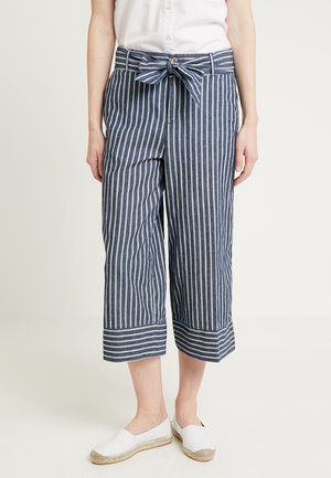 Mujer vistiendo pantalones cortos a rayas verticales azules y blancas con cinturón anudado, blusa blanca y zapatos de esparto blancos.