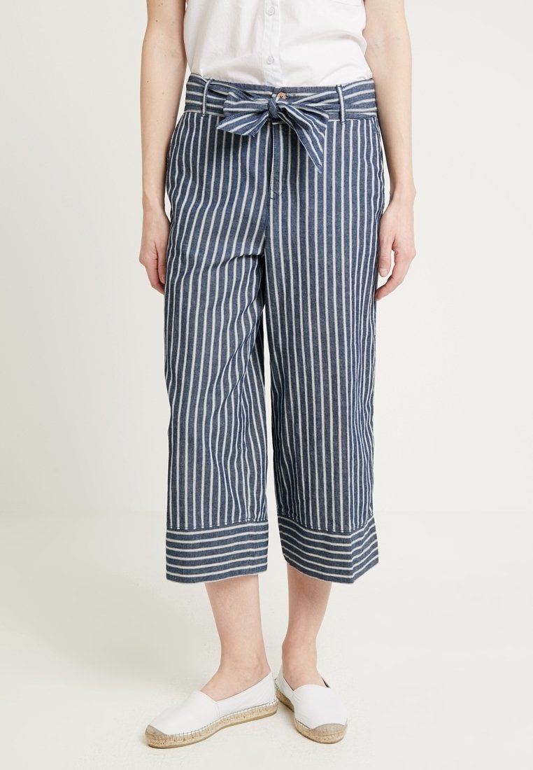 Nainen pukeutuneena sinivalkoraidallisiin, vyöllä solmittuihin lyhytlahkeisiin housuihin, valkoiseen yläosaan ja valkoisiin espadrillokengillä.