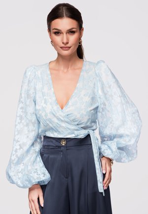 Femme portant un chemisier cache-cœur transparent bleu clair à motif floral avec des manches longues bouffantes et un pantalon sombre taille haute, posant avec une expression neutre.