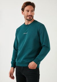 Teal színű, kerek nyakú pulóver, puha anyagból, bordás mandzsettával és szegéllyel; fehér hímzett logó található a mellkason. Fekete farmernadrág viselése.