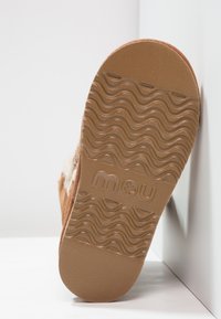 Semelle de pantoufle en suède marron avec un fond en caoutchouc texturé, présentant un motif ondulé et un logo de marque en relief, présenté sur un fond clair.