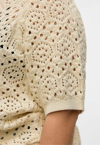 Top corto a maniche corte di colore beige in maglia, con intricati motivi in pizzo a forma circolare e a diamante sul tessuto, che mostra parte del braccio di chi lo indossa.