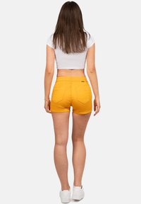Gele spijkerbroekshorts met een fitted ontwerp, vijf zakken en gestikte details, gecombineerd met een wit cropped top en witte sneakers.