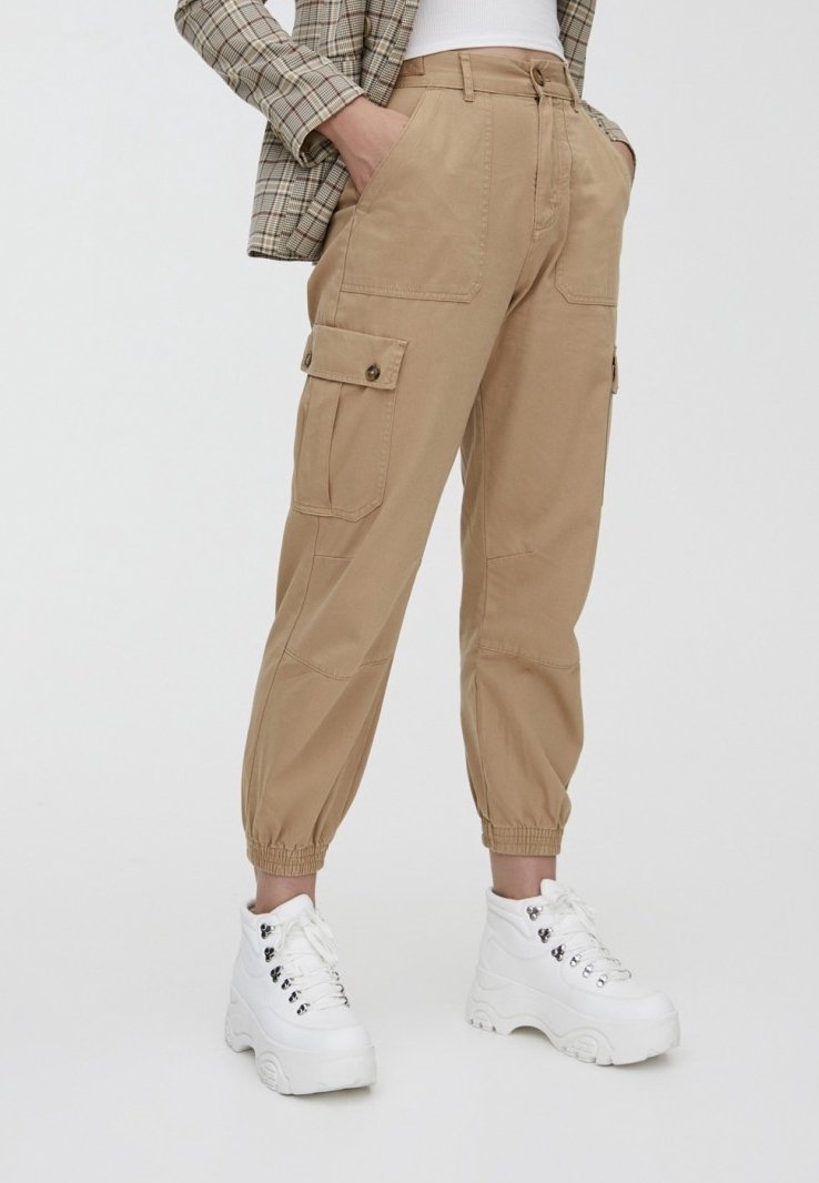 Pantalon cargo beige avec des poignets élastiques, des poches latérales et des détails en boutons, associé à des chaussures blanches imposantes. Tissu en coton, coupe décontractée.
