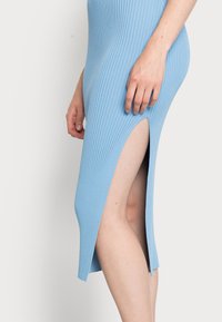 Vestido midi canelado de cor azul clara, com uma fenda lateral, silhueta ajustada e textura suave, revelando parcialmente a pele através da abertura.