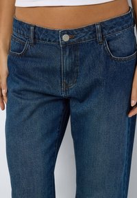 Jeans em denim azul de corte médio, com perna reta, apresentando cinco bolsos, fecho em botão prateado e ligeira descoloração no tecido.