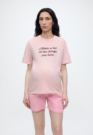 Frau mit langen dunklen Haaren trägt pinke Shorts und ein passendes pinkes T-Shirt mit der Aufschrift „Mach eine Liste der Dinge, die du liebst“ und steht vor einem weißen Hintergrund.