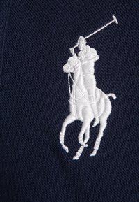 Marinblå tyg med en vit broderad polospelare på hästryggen, detaljerad med design av poloklubb och sadel.