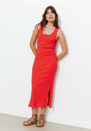 Femme en robe midi rouge sans manches avec des boutons sur le devant et une fente latérale, portant des sandales à imprimé léopard et un collier de coquillages, debout contre un mur blanc.