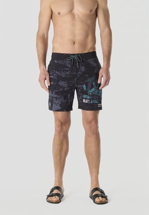 Hombre sin camisa con bañador negro estampado con calaveras abstractas en color azul verdoso y texto "BEATS & ROLL", de pie con sandalias negras sobre fondo gris.