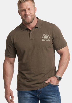 Uśmiechnięty umięśniony mężczyzna w brązowej koszulce polo z napisem „Mahalo & Waves Island Surf Co.” oraz w niebieskich jeansach, ręka w kieszeni, widoczny zegarek na nadgarstku.