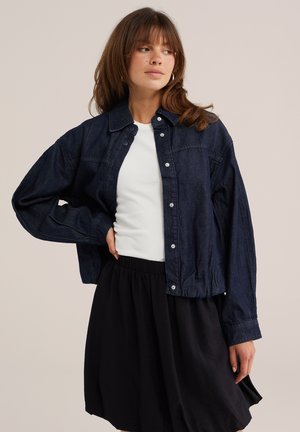 Donna con capelli castani che indossa una giacca di jeans blu scuro, una camicia bianca e una gonna nera, in piedi davanti a uno sfondo beige uniforme.