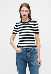 Γυναίκα με καστανά ίσια μαλλιά που φοράει κοντομάνικο πουκάμισο με οριζόντιες μαύρες και λευκές ρίγες και μπλε τζιν, με το ένα χέρι στην τσέπη.