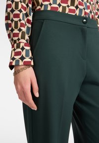 Linkerhand met een gouden schakelarmband, rustend naast donkergroene broeken en een multicolore geometrisch patroon manchette van een overhemd.