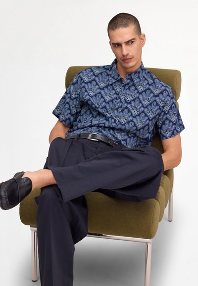 Homme assis sur une chaise vert olive, portant une chemise bleue à manches courtes à motifs, un pantalon noir et des mocassins noirs, les jambes croisées.