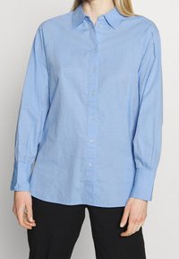 Femme portant une chemise boutonnée bleu clair à manches longues et un pantalon noir, se tenant contre un fond clair uniformément.