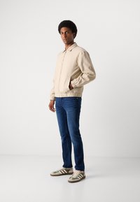 Beige Bomberjacke mit elastischen Bündchen, blaue Slim-Fit-Jeans und beigen Sneakers mit grünen Streifen. Einfarbiger Hintergrund, stehende Pose.