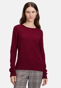 Kvinna iklädd en långärmad mörkröd tröja och grå rutiga byxor, stående med ett neutralt uttryck mot en enfärgad bakgrund.