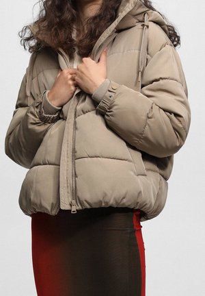 Personne portant une doudoune beige avec une capuche, partiellement zippée, et une jupe ajustée noire et rouge visible en dessous de la veste.