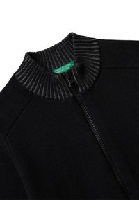 Sudadera negra con cremallera y un cuello acanalado oscuro texturizado. Presenta un cuerpo de tela suave y una etiqueta de marca verde en el interior del cuello.