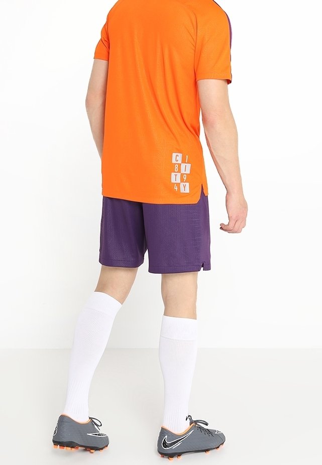 Pomarańczowa sportowa koszulka z fioletowymi akcentami, w połączeniu z fioletowymi spodenkami, białymi wysokimi skarpetami do kolan oraz szarymi korkami. Zawiera nadrukowane napisy.