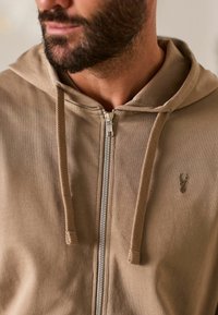 Lysbrun zip-up hættetrøje i stof, der har en kængurulomme, teksturerede snore til hætten og en subtil broderet accent.