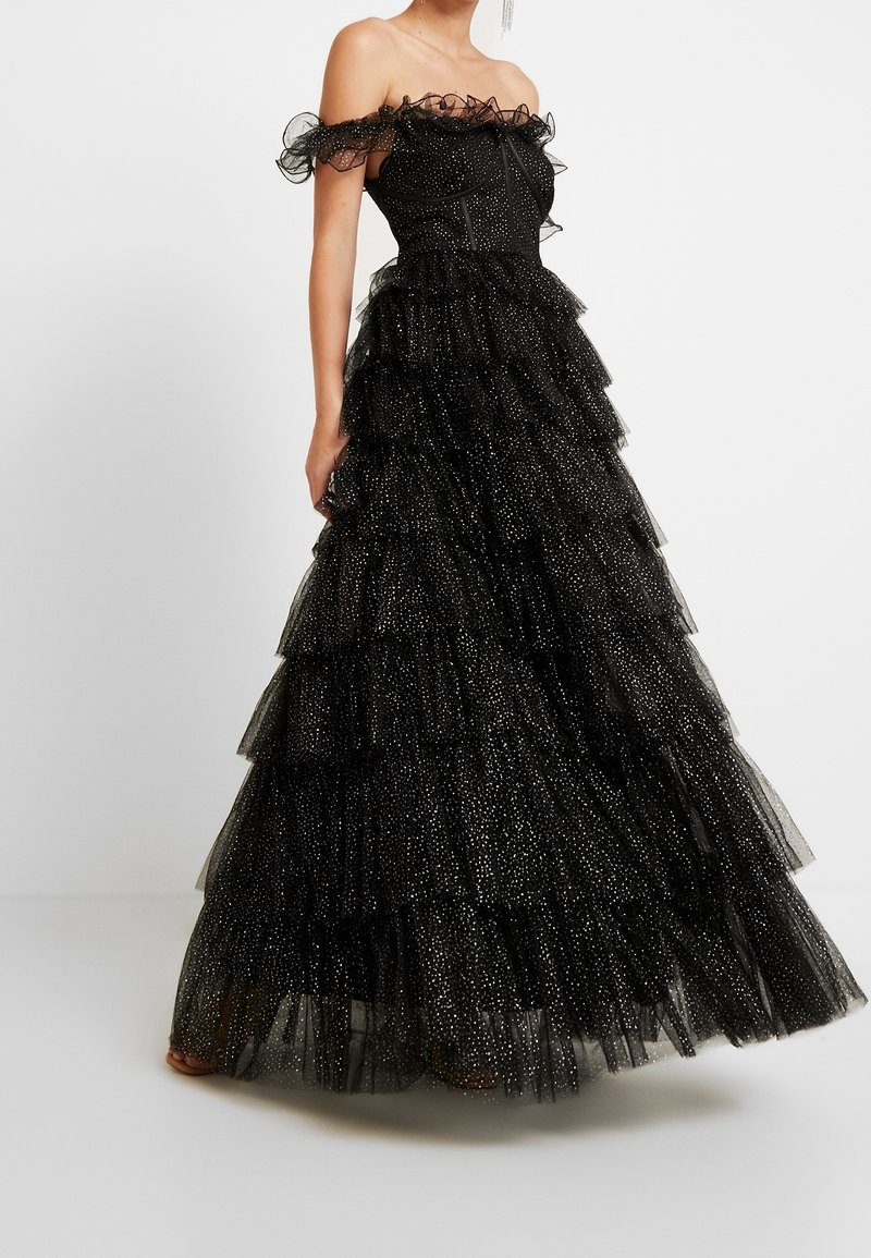 Femme portant une robe noire en tulle à épaules dénudées, à étages, ornée de pois brillants, debout sur un fond blanc uni.