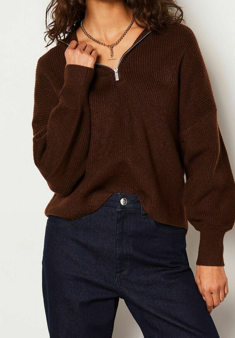 Femme portant un pull côtelé marron foncé avec un col à fermeture éclair argentée et un pantalon taille haute bleu foncé, tenant le bord du col.