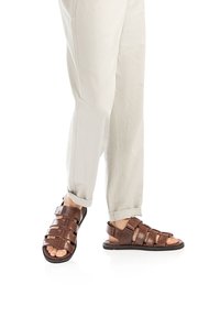 Braune Leder-Sandalen mit mehreren Riemen und Schnallenverschluss, ausgestattet mit einer flachen Sohle und offenem Zehenbereich, kombiniert mit hellen Hosen.