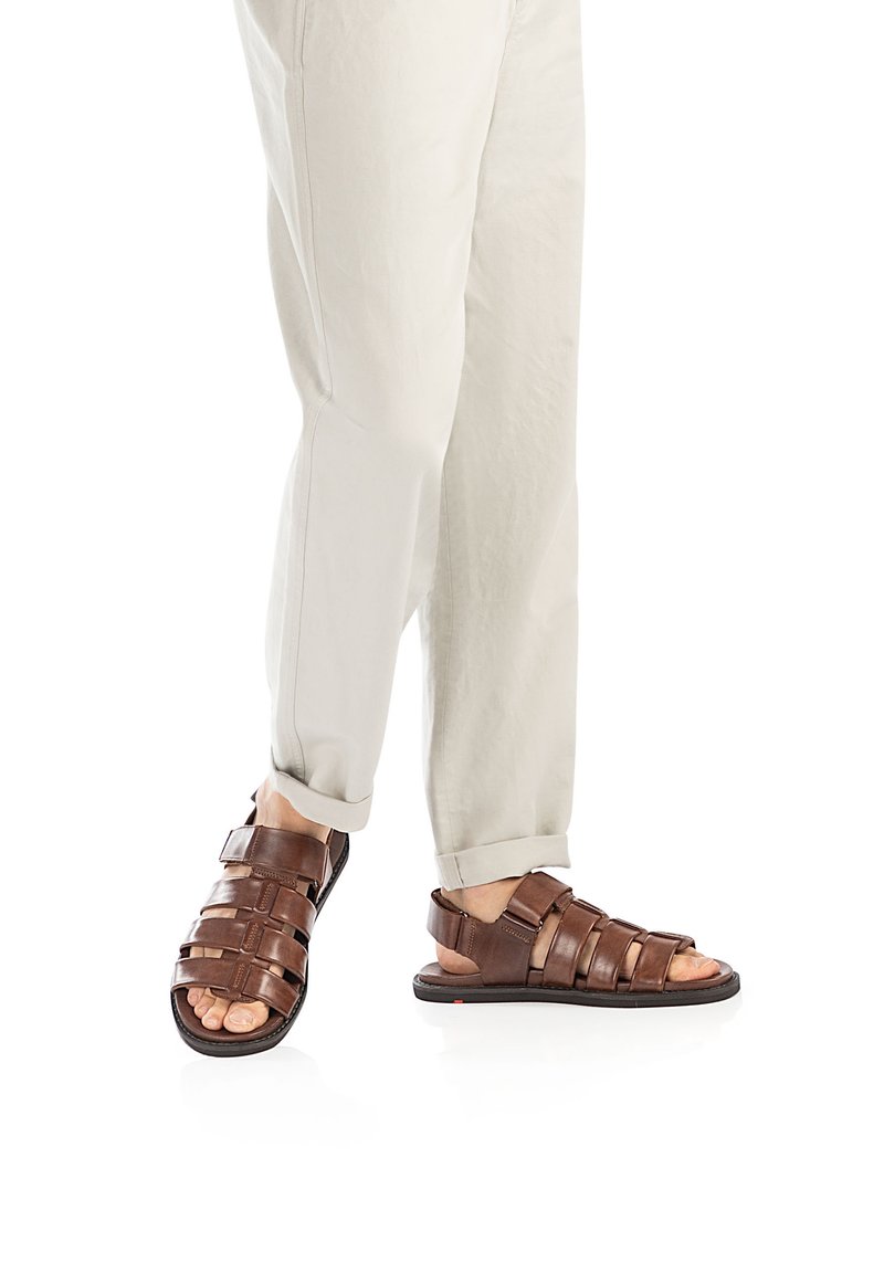 Braune Leder-Sandalen mit mehreren Riemen und Schnallenverschluss, ausgestattet mit einer flachen Sohle und offenem Zehenbereich, kombiniert mit hellen Hosen.