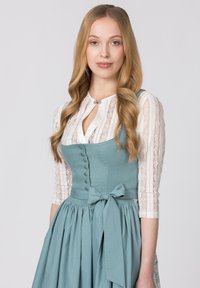 Traditionelles Dirndl in Teal mit einem knopfartigen Oberteil, gebundenem Taillenband und einem gepunkteten Rock. Die weiße Spitzenbluse hat vertikale Streifen.