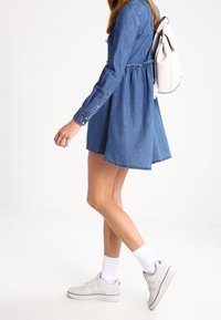 Denim-paita-mekko pitkillä hihoilla, kiinteällä vyötäröllä ja levenevällä hameella. Yhdistetty valkoisiin tennareihin ja vaaleansävyiseen reppuun.