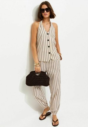 Donna che indossa un top smanicato beige e nero a righe verticali con bottoni e pantaloni abbinati, tiene in mano una borsa nera intrecciata, indossa occhiali da sole.