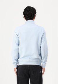 Sweatshirt azul claro com um colarinho alto, punhos e bainha canelados. O tecido parece suave, com uma textura lisa e sem padrões visíveis.